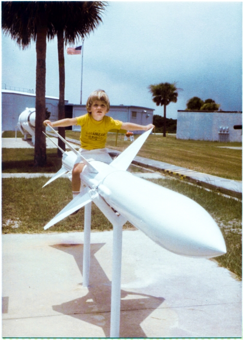 Kai MacLaren sits atop the Sparrow air-to-air missile at the Air Force Space and Missile Museum, Cape Canaveral Air Force Station, Florida. Partially visible behind him, extending out of frame on the left, is the blockhouse where launches at Complex 26 were conducted and controlled, and between it and Kai, a Minuteman intercontinental ballistic missile can also be seen. To the right of Kai, ground level, extending from out of frame right, all the way to the blockhouse, a metal-covered cable trench which carries electrical power and instrumentation cables to the launch pad can also be seen. Photograph by James MacLaren.